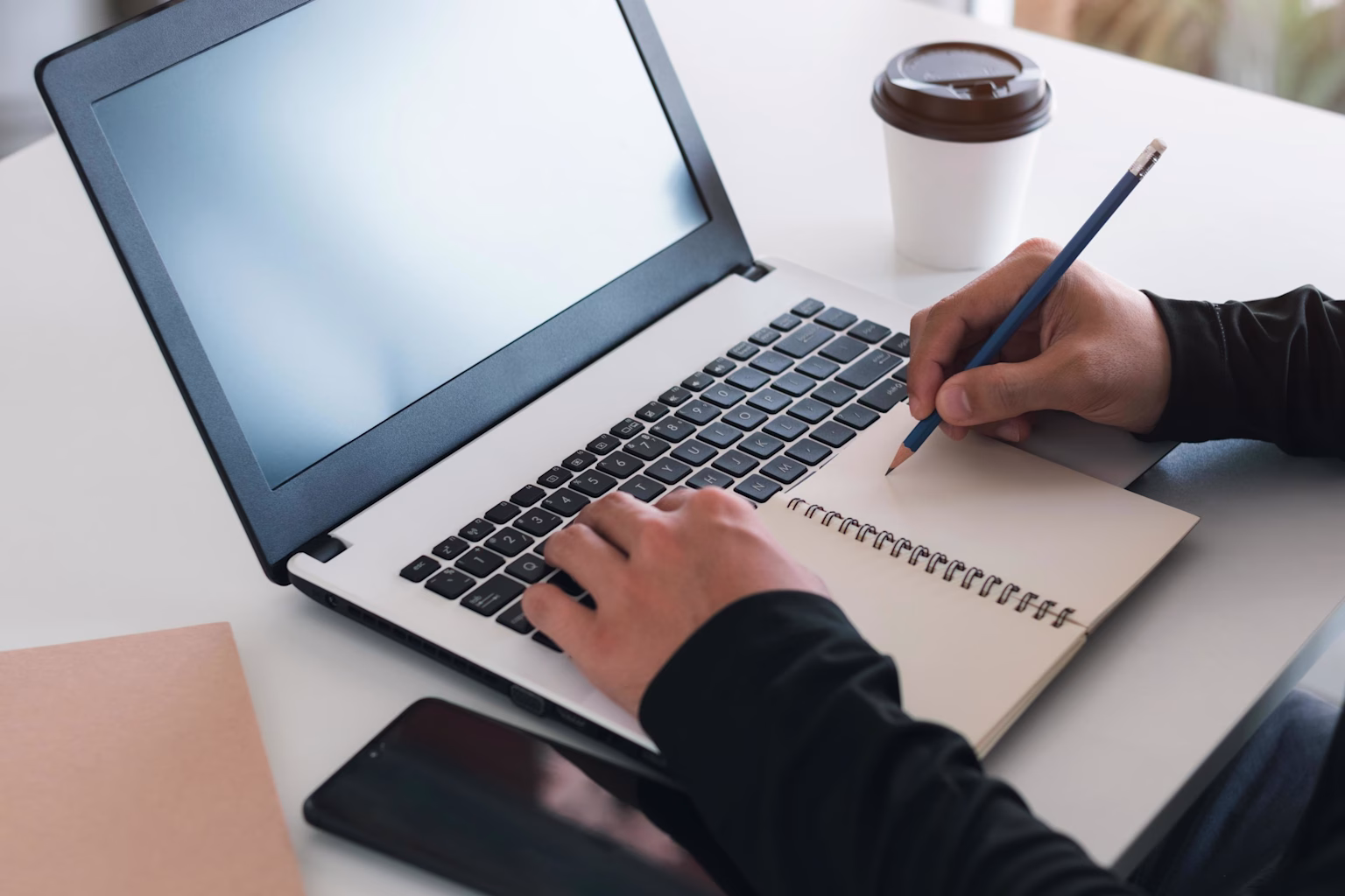 Person writing notes while studying with a laptop and coffee, symbolizing online learning or studying 